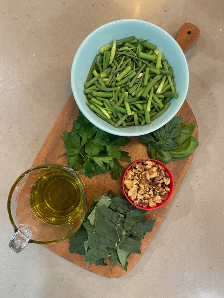 Fresh ingredients on a cutting board: garlic scapes, basil, parsley, walnuts, kale, and olives, ready to be transformed into a delicious homemade pesto sauce.