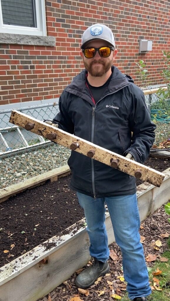 Efficient garlic planting: A man holds a specialized tool designed for quick garlic planting, resembling a dibbler, with dowels for creating six holes in the soil.